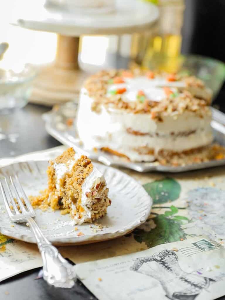 A slice of vegan carrot cake with cream cheese frosting rests on a white plate with a fork. The remaining cake is in the background, topped with frosting and walnuts. The table is decorated with illustrated papers.